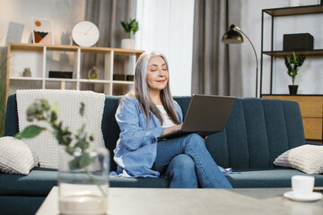 Cheerful mature caucasian woman sitting on couch and working on wireless laptop. Attractive...