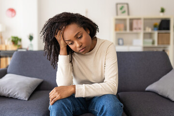 Depressed sad african american woman thinking about her problems while sitting alone on a sofa at home. Sadness and negative emotions concept.