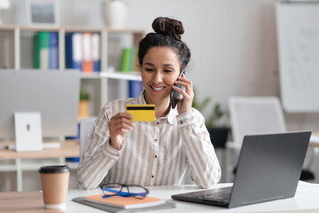 Cheerful woman working on laptop at office, holding credit card, calling her bank or webstore manager, using cellphone