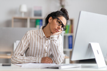 Portrait of bored businesswoman sitting at desk, leaning head on hand and looking at computer monitor