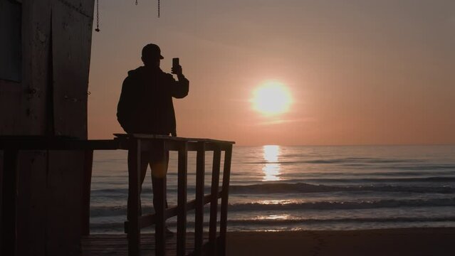 Silhouette of young man on observation deck shooting seaside sunset on phone. Yound adult male tourist makes a photo of sundown on clear sky, calm sea and empty beach. Slow motion.
