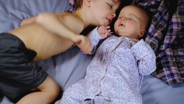 Older brother kissing newborn sister while lying on bed at home. Preschool boy kiss cheek little baby girl with love and care. Siblings playing together.