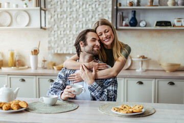 Glad young european lady hugging male with stubble drink coffee, enjoy free time in modern kitchen...