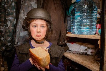 A sad child in a helmet hides in the basement from the shooting. A girl eats bread in a bomb shelter during the war in Ukraine.