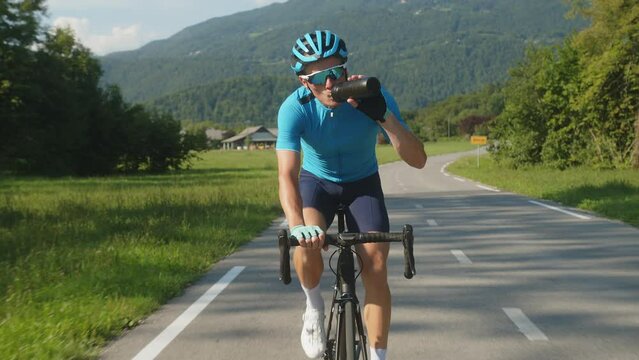 Professional male Caucasian racing cyclist drinking water from a sports bottle during training on an asphalt road, front view.