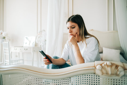 Portrait of young thoughtful woman using mobile phone, sitting on a bed and looking on a screen.
