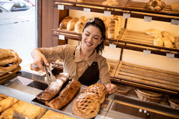 Deli worker in uniform working at supermarket bakery department selling pastry to the customers.