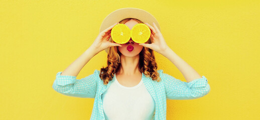 Summer portrait of cheerful young woman covering her eyes with slices of orange fruits and looking for something wearing straw hat on yellow background