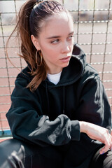 Close up outdoor portrait of stylish pretty woman with dark hair in black outfit is sitting on sport square and looking aside in sunlight