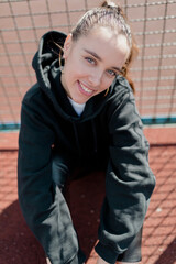 Wonderful pretty lady with dark hair wearing dark pullover is sitting on sport square and looking up into the camera with happy emotions