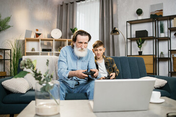 Smiling senior man grandfather and his little kid grandson in casual clothes using wireless joysticks for playing video games at home. Concept of retirement, people, fun and leisure time.