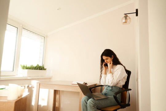 Distracted tired businesswoman feeling bored, looking at laptop chronic fatigue concept and speak by phone with colleagues at home. Online distane work and video business meeting.