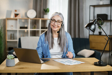 Attractive caucasian grey-haired woman sitting at desk and typing on laptop. Focused senior lady in denim shirt using portable computer for work at bright room at home.