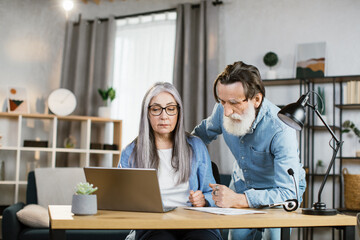 Senior caucasian man and woman in casual outfit looking at computer screen during working meeting. Mature business partners cooperating at bright office.