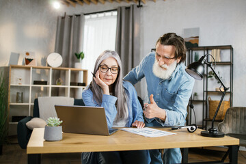 Fototapeta premium Beautiful senior man standing near caucasian woman that sitting at table and using laptop and showing thumb up. Two office workers in casual outfit. Work with modern technology.