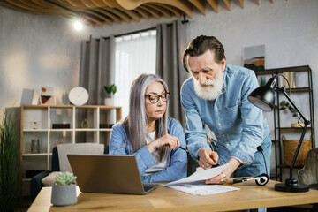 Obraz na płótnie Canvas Lovely aged man and woman analysing financial report while sitting at bright office. Pleasant old wife and her bearded husband using modern gadgets for work. Business and technology concept.