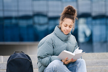 African american teenage student reading a book sitting on the steps of the university    