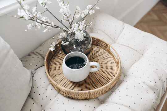 Spring breakfast still life composition. Cup of coffee and wickered tray on French mattress. Feminine styled photo. Floral scene with blurred white cherry tree blossoms. Bench near window. Selective