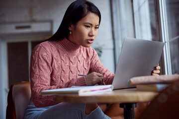 An asian female student is doing her assignment on a laptop while sitting in a coffee shop.