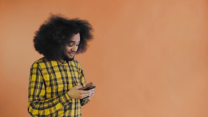 Fototapeta na wymiar A young man with an African hairstyle on an orange background takes out his phone, looks at the message and puts it back in his pocket. Emotions on a colored background
