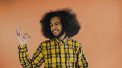 A young man with an African hairstyle on an orange background shows the OK gesture. Emotions on a colored background.