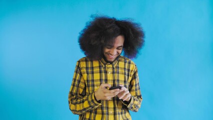 A young man with an African hairstyle on a blue background looks at the phone and happily texts with someone. Emotions on a colored background