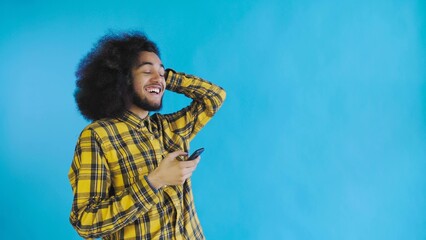 A young man with an African hairstyle on a blue background looks at the phone and is happily surprised. Emotions on a colored background