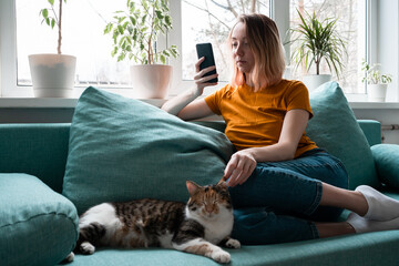 Young woman in yellow t-shirt sitting on sofa with cat holdind smartphone.