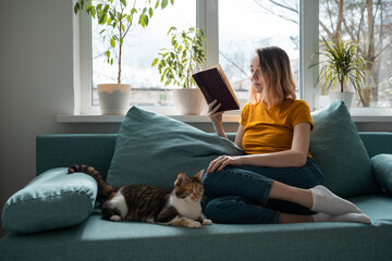 Young woman in yellow t-shirt reading book on sofa with cat.