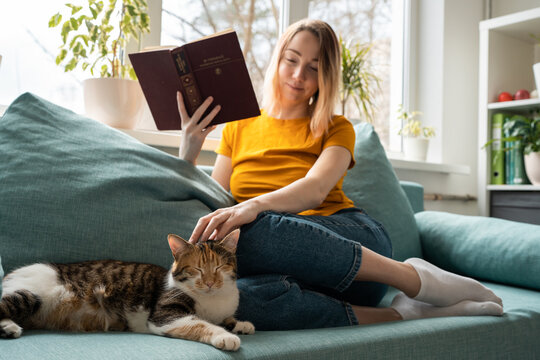 Young woman in yellow t-shirt reading book on sofa and petting cat.
