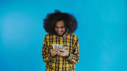 A happy African-American man holding banknotes in his hands, counting them and looking at the camera, standing isolated on a blue background