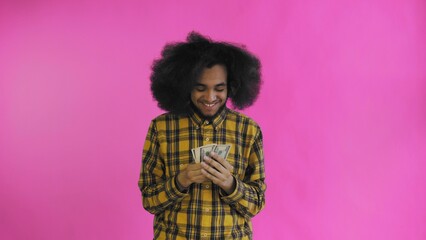 A happy African-American man holding banknotes in his hands, kissing them and looking at the camera, stands isolated on a pink background