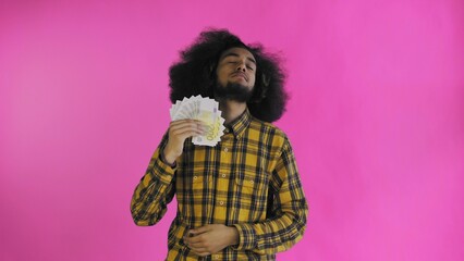 A happy African-American man holding a fan of banknotes in his hands and looking at the camera, standing isolated on a pink background