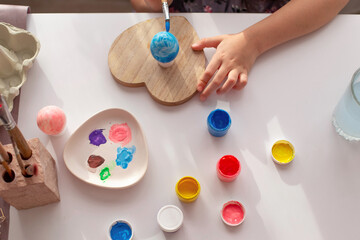 Kids hands paint an egg, on a white table with multi-colored paints in jars