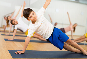 Fototapeta na wymiar Positive sporty teen boy doing yoga with sister and parents in fitness studio, standing in side plank or Vasisthasana pose..