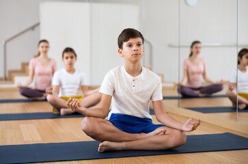 Concentrated teen boy sitting on mat in fitness center, making yoga meditation in lotus pose while...