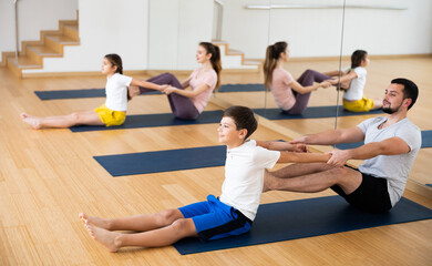 Fototapeta na wymiar Smiling teenage boy exercising with father at couple yoga class while his mother practicing with his sister, practicing stretching in pairs