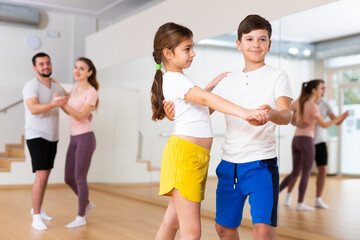 Young girl and boy dancing waltz together during group family training. Their parents dancing in background.