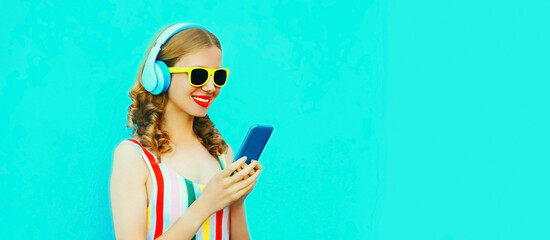 Portrait of happy smiling young woman listening to music in headphones with smartphone on blue...