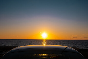 A Caribbean sun sets into the ocean and casts a pretty reflection on the roof of a car parked on the coastline. The romantic scene would be enjoyed by those in the vehicle