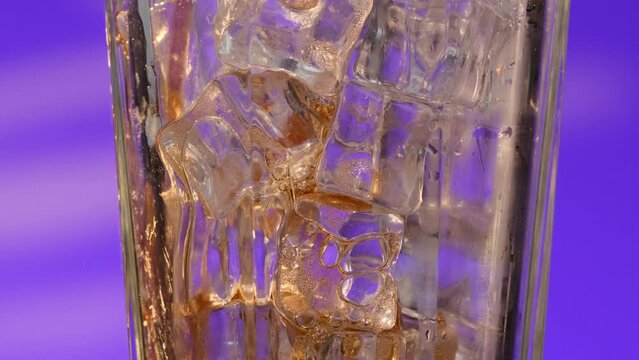 Close-up of a glass glass filled with ice on a purple background. The glass is filled with a carbonated drink passing through ice cubes. Bubbles rush up