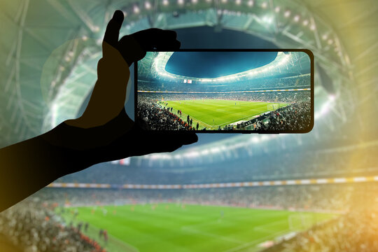 Fan hand with mobile phone photographing football game. Using a smartphone at a soccer stadium