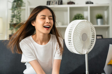Young smiling asian woman sitting on couch and turned on fan to cool herself with suffers from too...