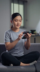 Asian woman sitting on sofa using smart phone and credit card for shopping online at home.