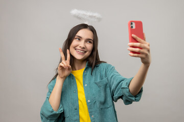 Portrait of friendly woman with nimb over head, holding cell phone, showing v sign to followers, livestream, wearing casual style jacket. Indoor studio shot isolated on gray background.