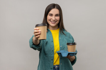 Portrait of friendly happy young adult woman offering coffee, giving drinks in disposable cups and smiling, wearing casual style jacket. Indoor studio shot isolated on gray background.