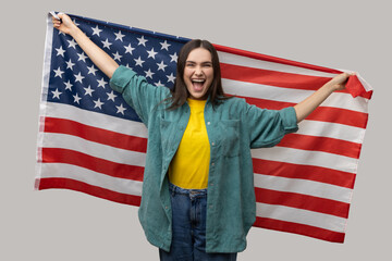 Happy positive woman dark hair standing with raised arms, holding USA flag, celebrating national holiday, rejoicing, wearing casual style jacket. Indoor studio shot isolated on gray background.