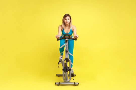 Attractive Confident Young Woman Riding On Spinning Bike, Cycling On Gym Machine Bicycle, Motivation And Sporty Goal Concept, Wearing Blue Sportswear. Indoor Studio Shot Isolated On Yellow Background.