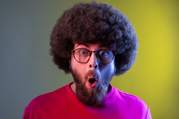 Fototapeta na wymiar Hipster man with Afro hairstyle standing with mouth open in surprise, has shocked expression, unbelievable news, wearing red sweatshirt. Indoor studio shot isolated on colorful neon light background.