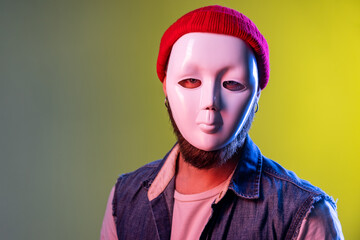 Unknown anonymous hipster man in white mask on face, hiding personality, conspiracy and privacy, secrets, wearing hat and denim vest. Indoor studio shot isolated on colorful neon light background.